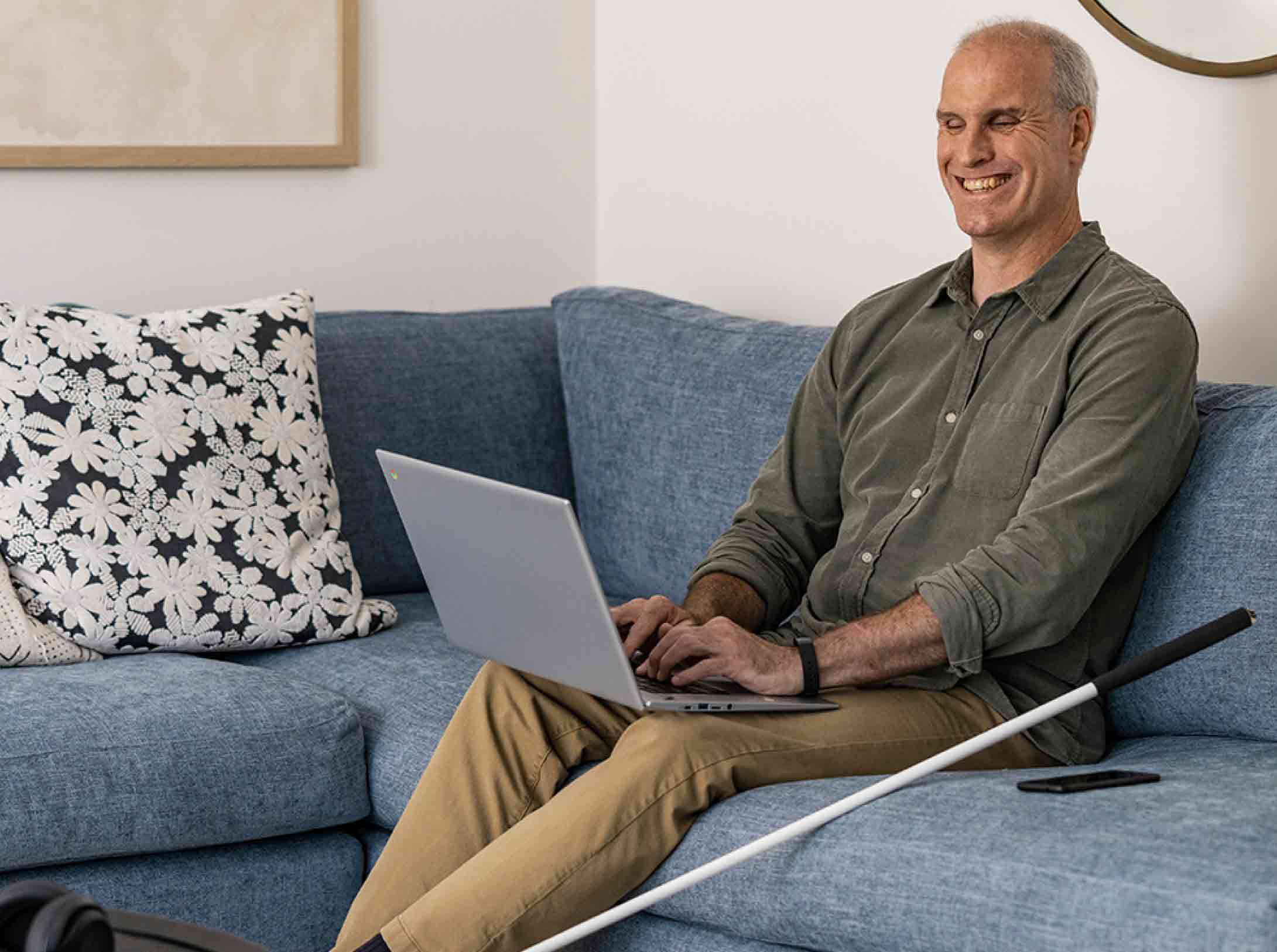 Una persona più grande è seduta su un divano blu e digita su un Chromebook aperto tenuto sulle gambe con un bastone bianco per non vedenti appoggiato alla sua sinistra.