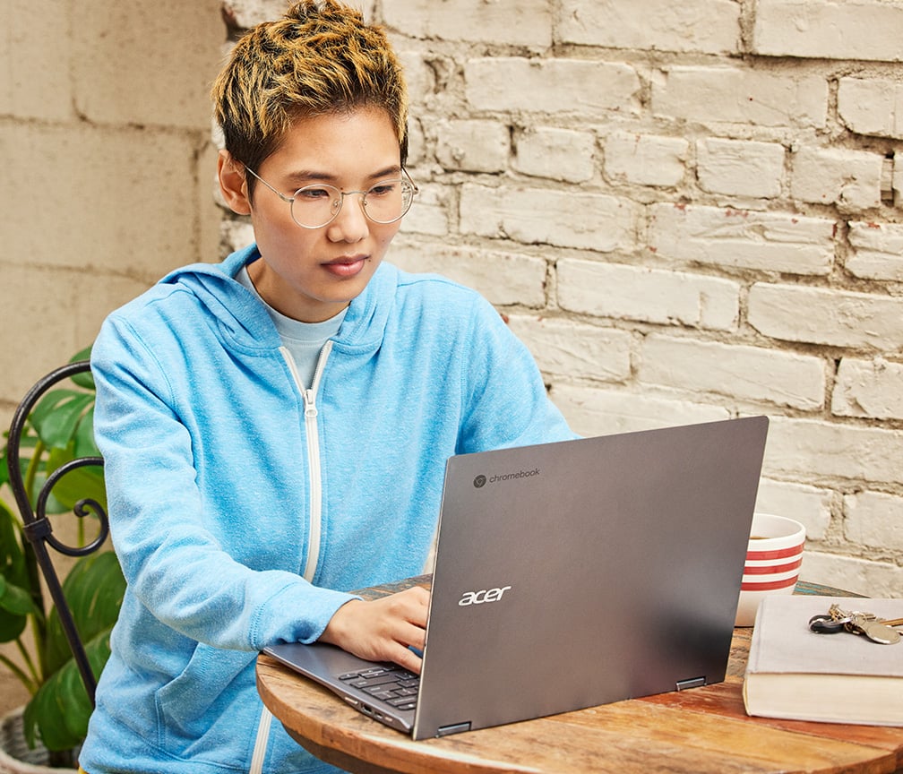 Uma pessoa com camisa listrada está sentada em uma mesa e trabalha em um Chromebook conectado a um monitor externo.