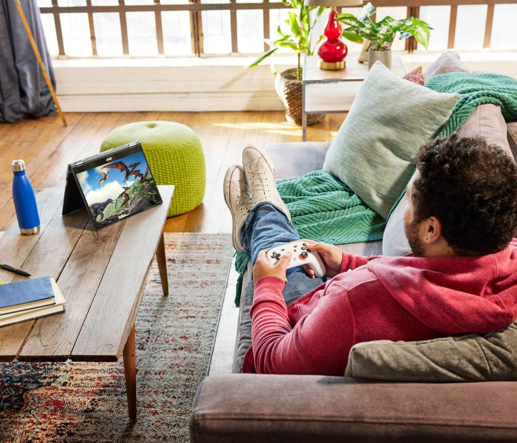 A person sits on a couch with their feet up and plays video games on a Chromebook using a game controller.